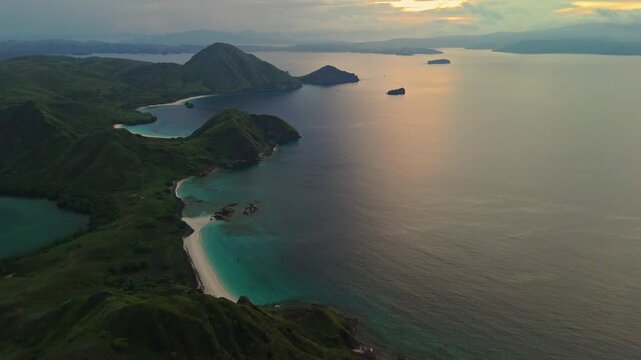 Drone pullback shot at sunrise over Padar Island in Komodo National Park, Indonesia, showing lush green hills during rainy season, turquoise bays, white beaches and calm blue water.