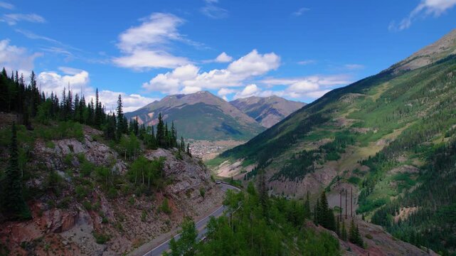 Aerial reveal of historic Colorado mining town, Silverton, on a bright summer day with lush, green forests and towering peaks all around. Cars drive along the steep winding road to the mountain town