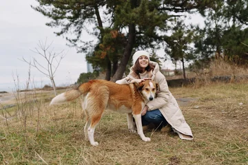 Fotobehang Onderzeeër Woman kneeling beside a large dog in an autumnal park, wearing a warm coat and hat, smiling as they bond in an open field with trees and overcast sky together.  © SHOTPRIME STUDIO