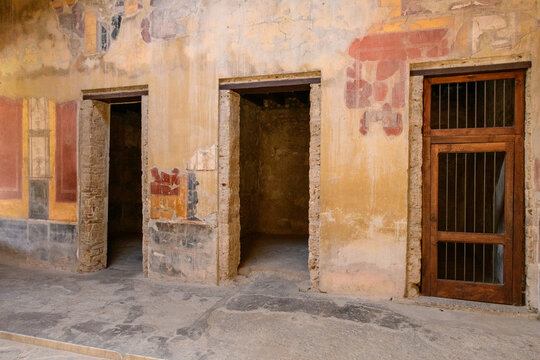 Ancient stone doorways and a wooden gate line a faded frescoed wall in the House of Menander, Pompeii, Italy. Cracked plaster, worn masonry, and remnants of classical wall painting evoke the