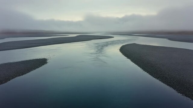 Winding glacial river flowing through dark gravel plain under a dramatic foggy overcast sky in a remote desolate landscape.