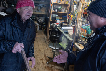 Two men measuring wooden board with tape measure inside garage workspace focused on material length and preparation stage.