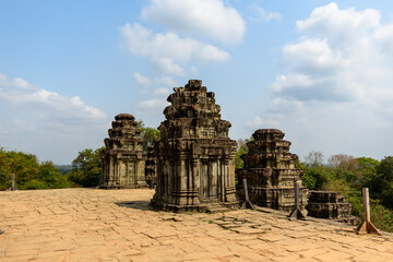 Obraz premium Three weathered sandstone towers rise from the sunlit terrace of Phnom Bakheng temple near Siem Reap, Cambodia. The ancient structures stand against a backdrop of green treetops and a sky dotted with