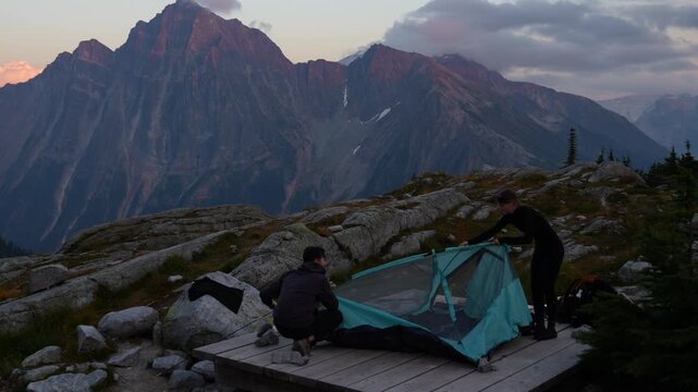 Couple sets up camping tent on a rocky slope with mountain ranges behind them at dusk in Glacier National Park, British Columbia. Real-time outdoor footage.