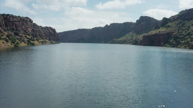 Low-altitude drone captures Pennar River reservoir stretching between Gandikota Canyon's towering red rock walls and green canyon rims under partly cloudy skies.