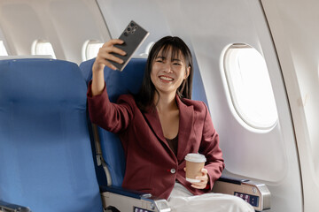 Fototapeta premium A young woman sits by the airplane window holding a smartphone and a cup of coffee, enjoying a quiet moment, modern travel comfort, and relaxed personal time during a smooth flight.