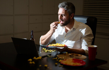 Businessman working late at office desk. Employee after night work. Overtime at night office. Freelancer finishing project on laptop after hours. Overworked man eating during night work. Night job.