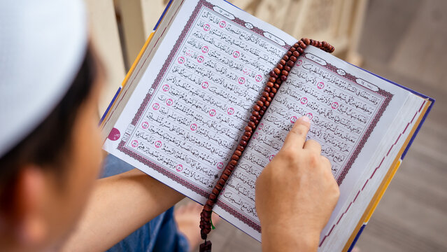 A close-up overhead view of a finger pointing at Arabic script in the open holy Quran during recitation (tilawah), with wooden prayer beads draped over the page.