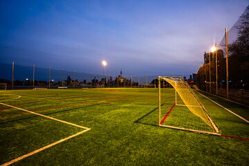 Fototapeta premium Empty stadium field with football lines and Manhattan skyline in the background. Soccer stadium in NYC with green turf and city lights at dusk. Urban football stadium overlooking Manhattan.