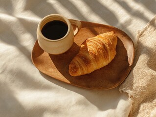 A cup of black coffee sits on a wooden tray with a golden croissant, bathed in warm sunlight and shadow.