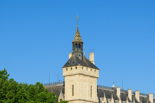 Elegant stone clock tower with a pointed spire and intricate rooftop details rises above lush green trees and classic rooftops, set against a vivid blue sky in central Paris.