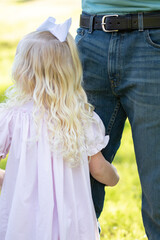 Young blonde girl wearing light pink dress hugs father's leg in outdoor park setting