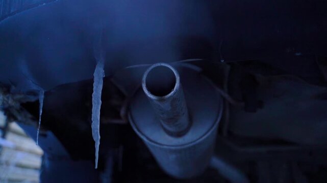 An icicle hangs on the rear bumper of a car against the background of the exhaust pipe while the car is warming up. Frosty morning