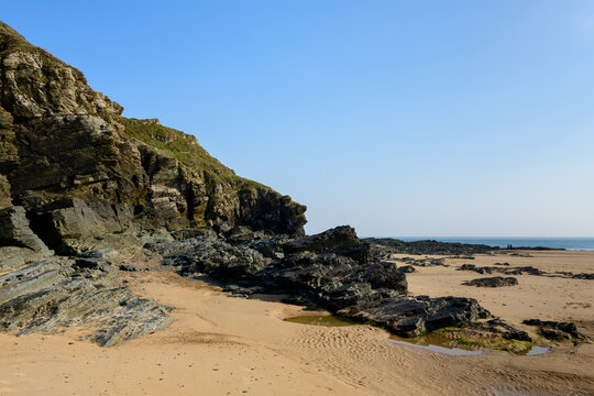 Sharp, dark rock formations jut from a quiet sandy beach at Cap de Carteret, with clear blue sky and gentle sunlight creating strong contrasts and natural textures.