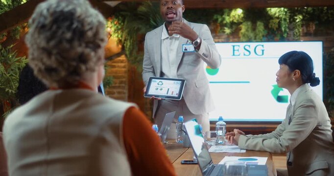 Open communication during ESG meeting as Black leader presents data on tablet while multiracial team listens and analyzes results, supporting decision making and strategic discussion in eco office.