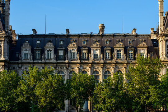 Symmetrical upper facade of the Hotel de Ville in Paris, adorned with sculpted stonework and statues, rises above leafy green trees in warm evening sunlight against a clear blue sky.