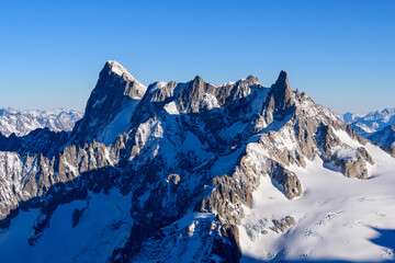 Obraz premium The dramatic, jagged summits of the Grandes Jorasses rise steeply above pristine glaciers and rugged rock faces, illuminated by crisp morning light under a cloudless blue sky in the French Alps.