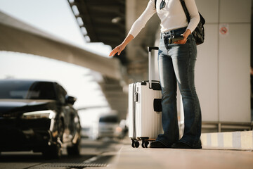 Fototapeta premium Young Asian woman traveler standing at an airport curbside with a rolling suitcase and smartphone, waiting for pickup or transportation, representing modern travel and urban mobility.