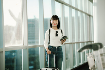Fototapeta premium Young Asian woman standing in a modern airport terminal, using a smartphone while holding a passport, with travel luggage nearby, preparing for a journey in a bright, contemporary space.