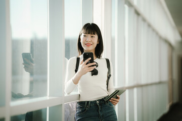 Fototapeta premium Young Asian woman standing in a modern airport terminal, using a smartphone while holding a passport, with travel luggage nearby, preparing for a journey in a bright, contemporary space.