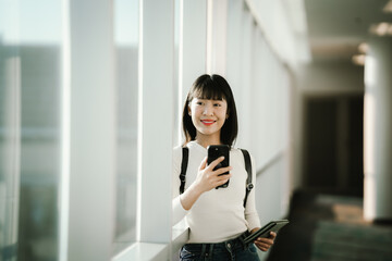 Fototapeta premium Young Asian woman standing in a modern airport terminal, using a smartphone while holding a passport, with travel luggage nearby, preparing for a journey in a bright, contemporary space.