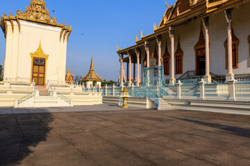 Naklejka premium Expansive view of the Royal Palace courtyard in Phnom Penh, Cambodia, featuring ornate Khmer architecture, golden spires, and decorative gates under clear blue sky. The scene is bathed in warm