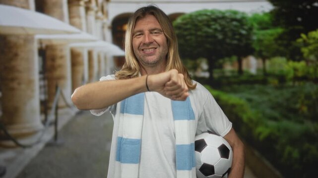 Man with long blond hair wearing white shirt and blue scarf holding soccer ball raises fist in campus cloister; joy strength.
