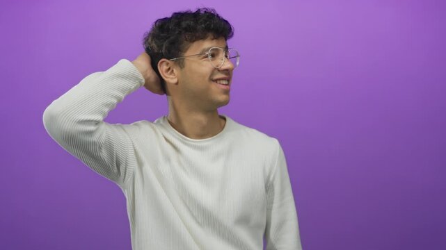 Man wearing clear glasses lightly touches the back of his head while smiling against a purple studio wall; shyness uncertainty awkwardness hesitation.
