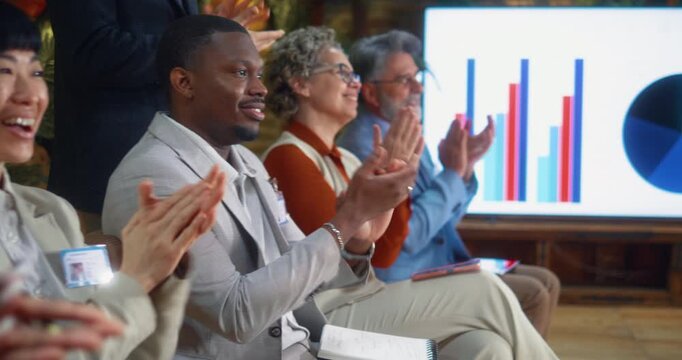 Multiracial business people seated in a row applauding after a seminar or meeting, celebrating positive results, engagement, and shared success in a modern corporate training setting.