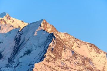 Fototapeta premium Golden sunlight illuminates the snowy, rugged slopes of Aiguille de Bionnassay, revealing crisp textures and sharp contrasts beneath a clear blue sky in the French Alps.