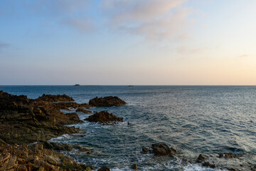 Jagged rocks emerge from the shoreline into the calm sea at Cap de la Hague, illuminated by soft evening light and a pastel sky. The tranquil scene highlights rugged textures and the serene expanse of