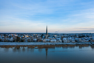 Fototapeta premium A tranquil winter scene shows the snow-dusted rooftops of Sully sur Loire, dominated by a tall church spire, reflected in the calm waters of the Loire River under a pastel evening sky.