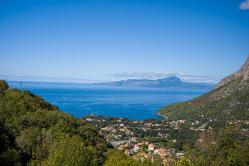 Obraz premium Elevated view of a coastal settlement nestled among lush green hills with sweeping vistas of the Tyrrhenian Sea and distant mountains under a clear blue sky. Vibrant sunlight enhances the vivid colors