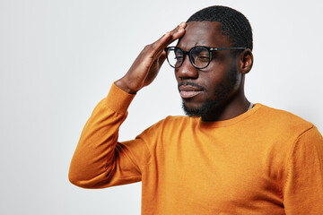 Fototapeta premium African American man with dark skin wearing glasses and orange sweater touching forehead with closed eyes, showing stress or headache on white background.