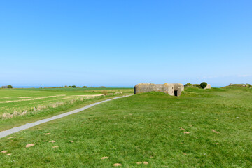 Fototapeta premium Historic concrete bunker stands on a green headland overlooking the sea at Longues-sur-Mer, Normandy. The open landscape and clear blue sky create a serene, expansive atmosphere.