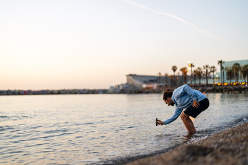 Young man capturing the beautiful sunset over the city beach while standing by the calm ocean water