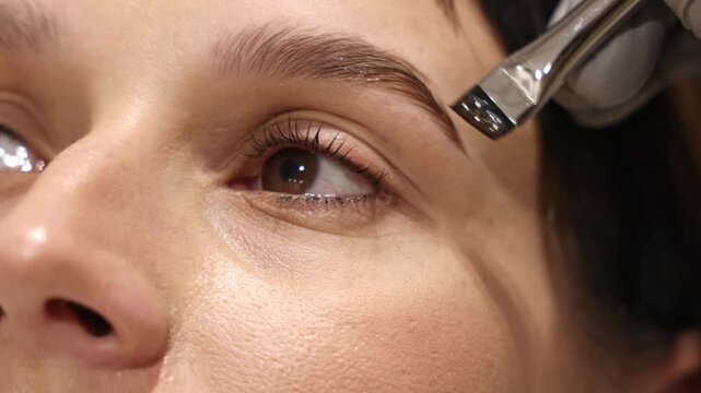 Woman during eyelash and eyebrow lamination in beauty salon, showing steps of foam application, brushing, tinting and styling. Close up of female eye, professional cosmetic treatment.