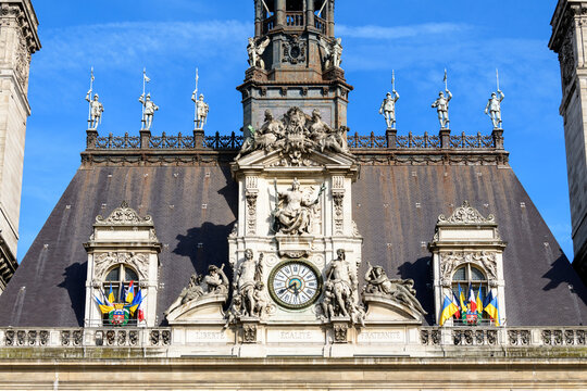 Elaborate stone statues and a decorative clock adorn the steep slate roof of Hotel de Ville in Paris, with intricate ironwork and vibrant flags under a clear blue sky.