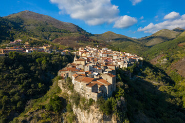 Obraz premium Aerial view of Tortora, Italy, showing a cluster of old stone houses with terracotta roofs dramatically situated on a rugged cliff, surrounded by green hills and distant mountains under a vibrant blue