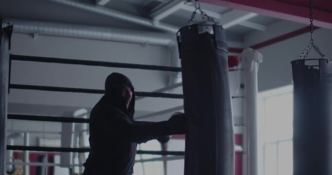 Male boxer training with punching bag in a boxing gym. Intense punching bag session as a man boxer sharpens power and technique in shadows. Self defense concept. Slow motion 4k video.
