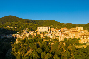 Obraz premium Historic Pisciotta sits atop a sunlit hillside, surrounded by dense green forest and rolling mountains under a clear blue morning sky. Warm golden light highlights the rustic architecture and natural