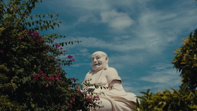 Laughing Buddha statue in Thailand framed by vibrant trees and flowers under a bright blue sky.