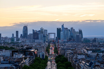 Obraz premium Wide view of La Defense business district rising above Paris rooftops, with glass skyscrapers and the Grande Arche silhouetted against a glowing evening sky.