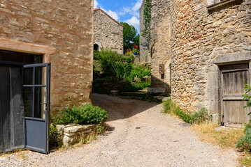 Fototapeta premium Quiet gravel lane in La Couvertoirade bordered by textured stone buildings, open blue-framed door, weathered wooden doorway, and lush greenery under a bright summer sky.