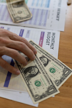Hand placing US dollar cash on monthly budget worksheet on wooden desk. Concept of personal finance, budgeting, expense tracking, saving money, household accounting and financial planning.