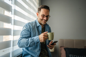 Man relaxing at home, using smart phone, drinking coffee