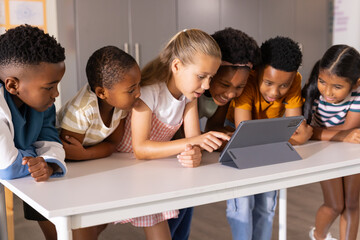 Diverse school-age students leaning over tablet in folio case and stand on white table in classroom