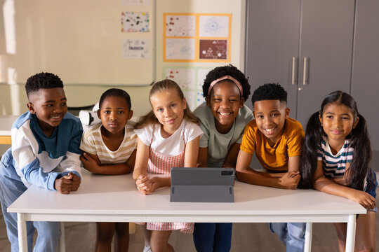 Diverse children leaning on white rectangular table watching tablet on folio stand in classroom