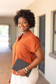 Adult African woman standing wearing burnt-orange shirt while holding tablet in covered corridor