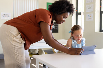 African American teacher helping school-age girl viewing tablet in blue folio case in classroom